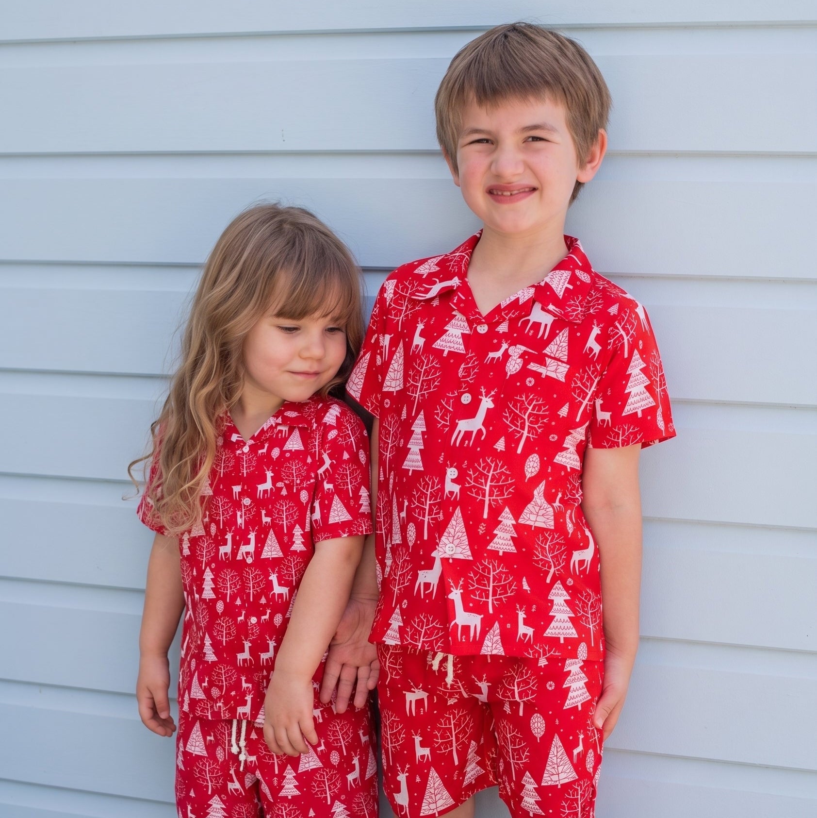 Two children wearing matching red holiday pajama sets with a white Christmas tree and reindeer print, standing barefoot against a light blue siding wall.