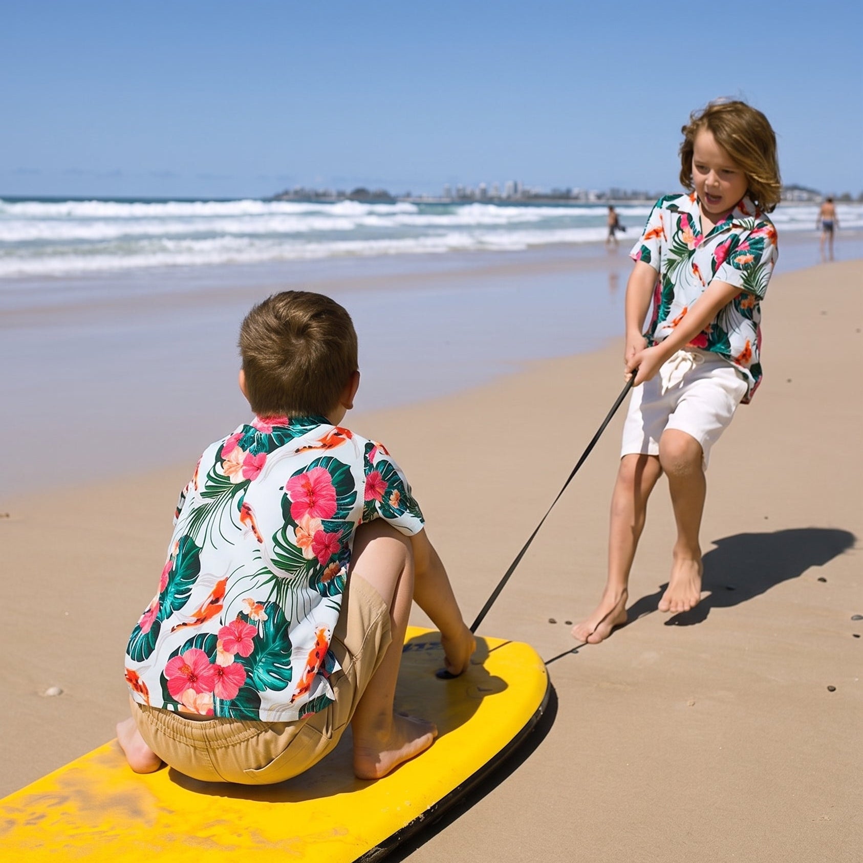 Two children on a sandy beach, one sitting on a yellow surfboard pulling another child with a rope. Both are wearing matching colorful tropical floral shirts and shorts, with the ocean and blue sky in the background.