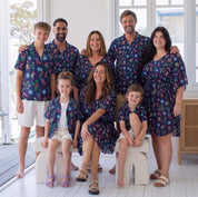 A family of seven posing together in matching dark blue tropical print swim shirts and swim shorts on a white-decked patio in front of a bright house.