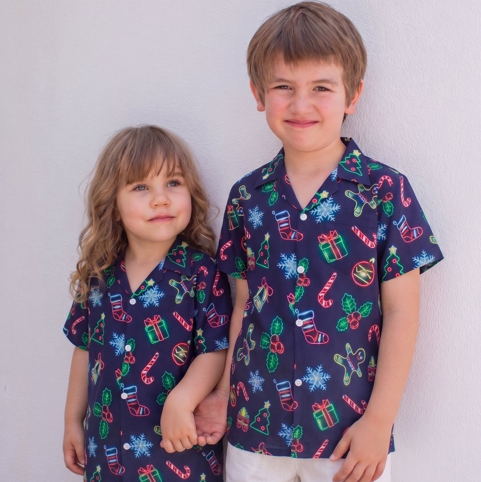 Two children, a boy and a girl, standing side-by-side and holding hands. They are wearing matching navy blue short-sleeve button-up shirts with a colorful Christmas-themed print of candy canes, snowflakes, ornaments, and gift boxes, paired with white shorts, against a plain white wall.