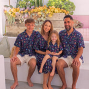 A family of four wearing matching navy blue paisley Hawaiian shirts and shorts sets, sitting together on a white outdoor bench with a colorful flower box in the background.