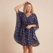A woman modeling a dark navy blue kaftan dress with a festive multicolor holiday print of candy canes, snowflakes, and gifts, set against a solid beige background.