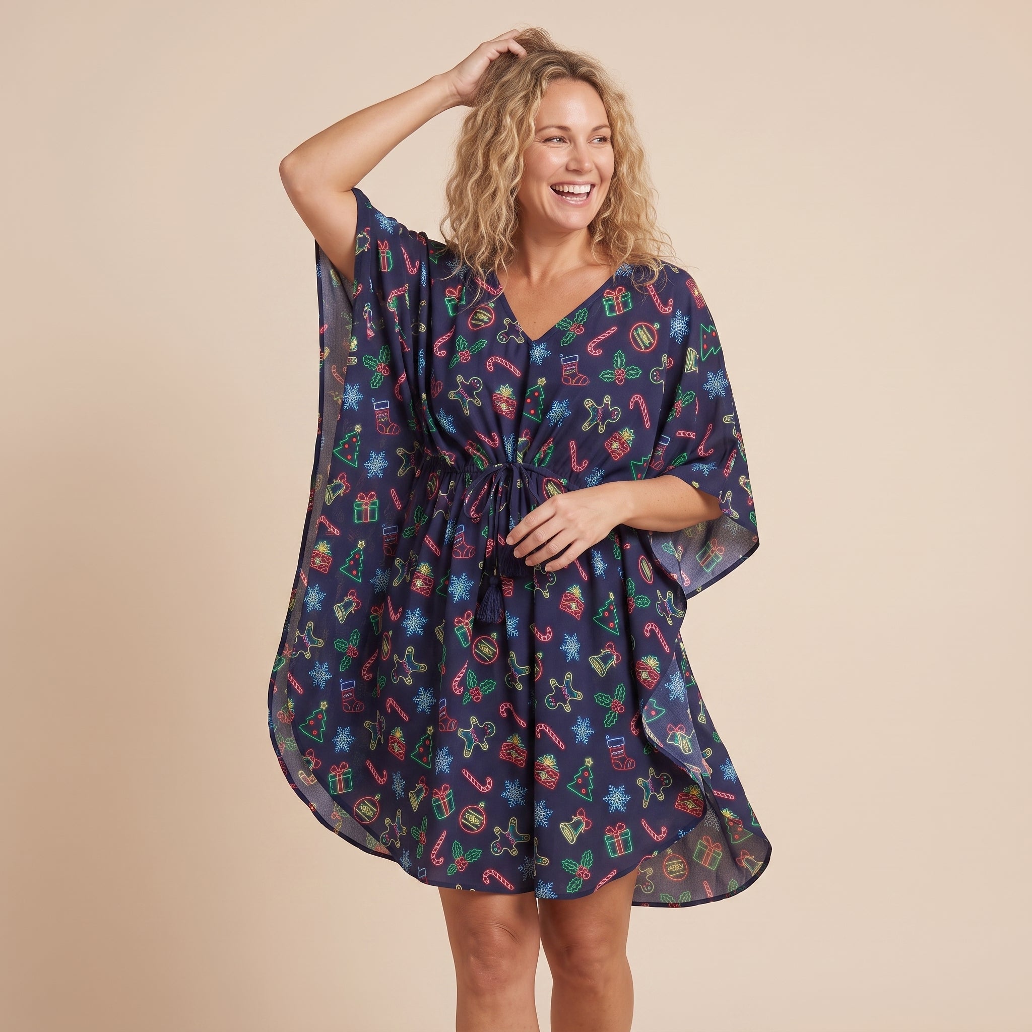 A woman modeling a dark navy blue kaftan dress with a festive multicolor holiday print of candy canes, snowflakes, and gifts, set against a solid beige background.