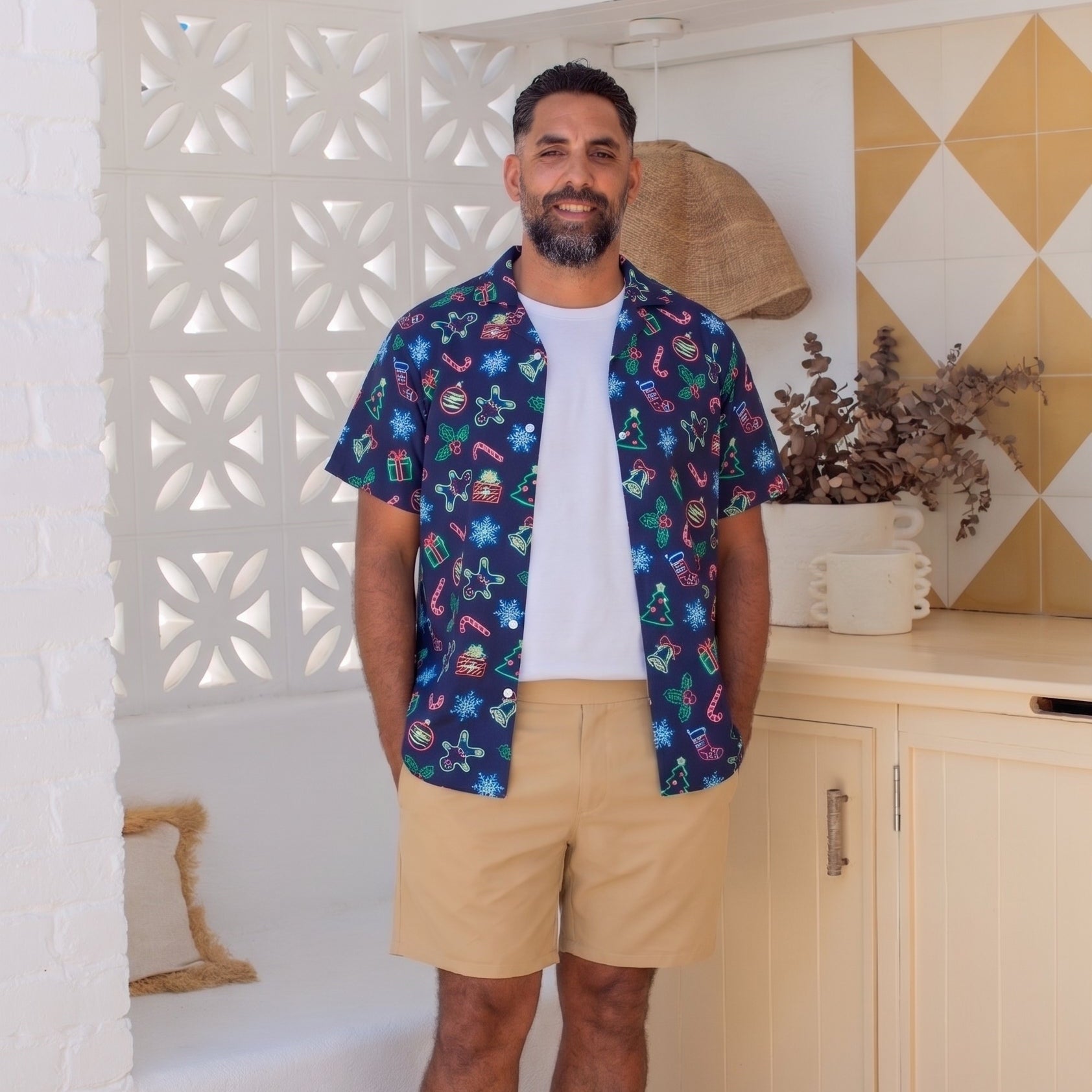 A man stands barefoot in a bright, modern room wearing a short-sleeve button-up shirt with a colorful tropical print, a white t-shirt, and khaki shorts.