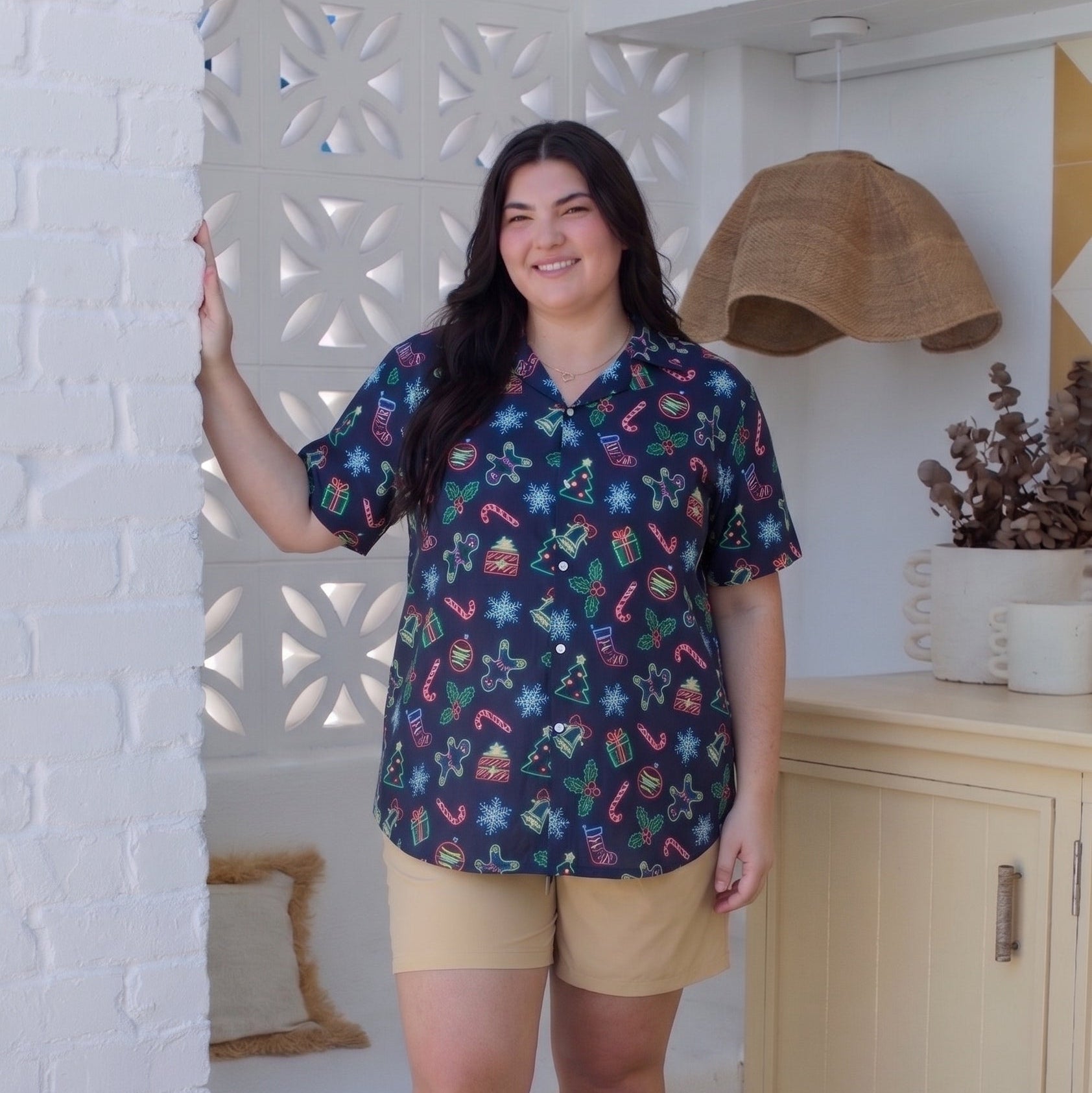 A woman stands in a bright indoor setting wearing a short-sleeve button-up shirt with a colorful holiday print, tan shorts, and black sandals.
