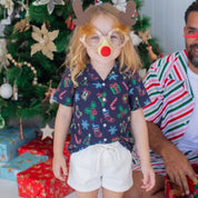 A young girl wearing a Christmas-themed novelty eyeglasses and antler headband with a red nose stands in front of a decorated Christmas tree, with a man in a striped shirt sitting beside her.