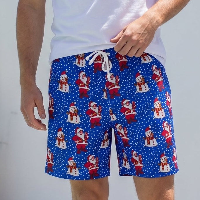 A man wearing blue swim trunks with a red and white Santa Claus pattern, a white t-shirt, and brown sandals, standing on a wooden deck next to a pool.