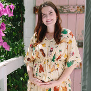 A pregnant woman wearing a short-sleeve, drawstring babydoll dress with a colorful cartoon animal print, standing on a porch in front of a pink wooden door.