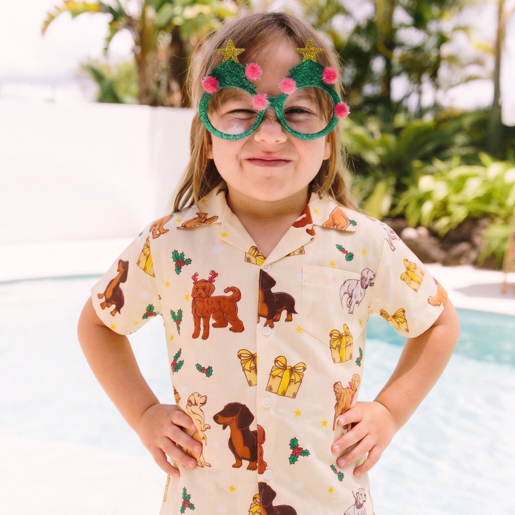 A young girl stands in front of a swimming pool wearing a short-sleeve button-up shirt with a dog print, khaki shorts, and novelty green glasses with pink flowers.