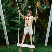 A young boy stands barefoot on a white rope swing in a lush garden, wearing a cream short-sleeve t-shirt with a colorful dog print and light beige shorts.