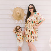 A woman and a child wearing matching cream-colored short-sleeve dresses with a colorful woodland animal print, standing against a white siding wall with a straw hat hanging on the wall.