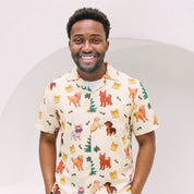 A smiling man wearing a short-sleeve button-up shirt with a cream base and a colorful print of various dog breeds, Christmas trees, and gift boxes, set against a minimalist white background.