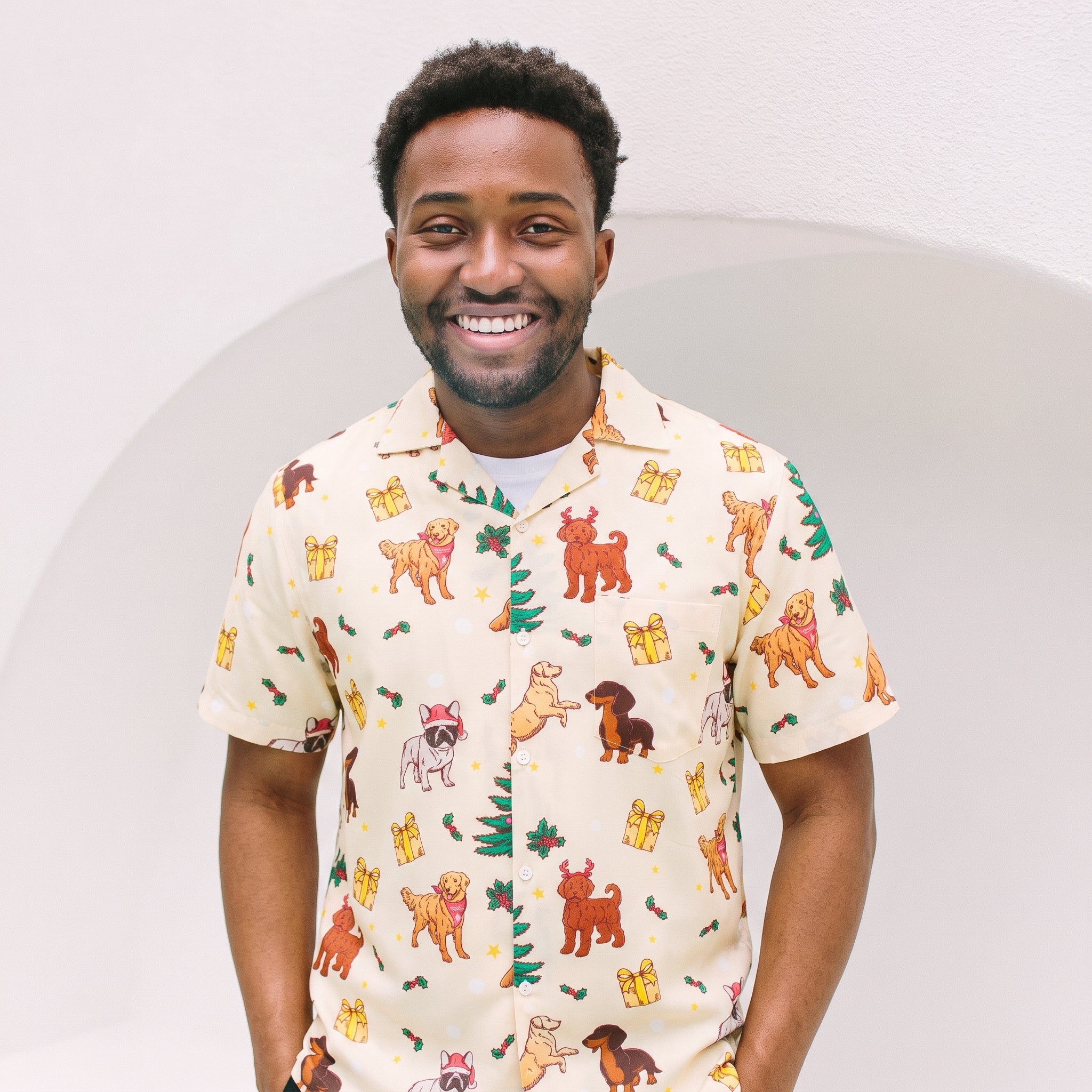 A smiling man wearing a short-sleeve button-up shirt with a cream base and a colorful print of various dog breeds, Christmas trees, and gift boxes, set against a minimalist white background.