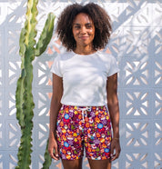 A woman stands outdoors in front of a white geometric wall, wearing a white t-shirt and colorful patterned shorts. A tall green cactus is visible to her left.