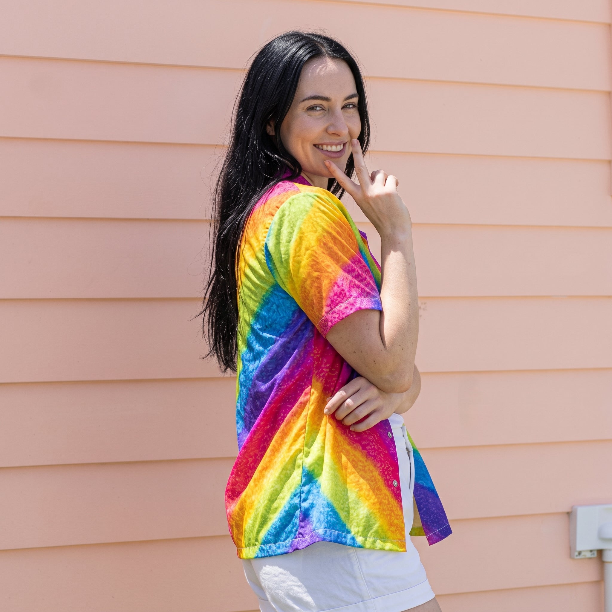 A woman with long black hair stands in front of a peach-colored horizontal siding wall, wearing a short-sleeve rainbow tie-dye shirt and white shorts.
