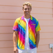 A smiling man wearing a vibrant rainbow-striped short-sleeve button-up shirt over a white t-shirt and white shorts, standing against a peach-colored house siding background.
