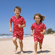 Two children running on a sandy beach wearing matching red short-sleeve camp shirts and shorts sets with a white tropical pattern.