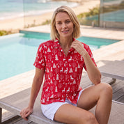 A woman sitting on a poolside lounge chair wearing a red short-sleeve button-up shirt with a white Christmas tree and deer pattern, paired with white shorts.