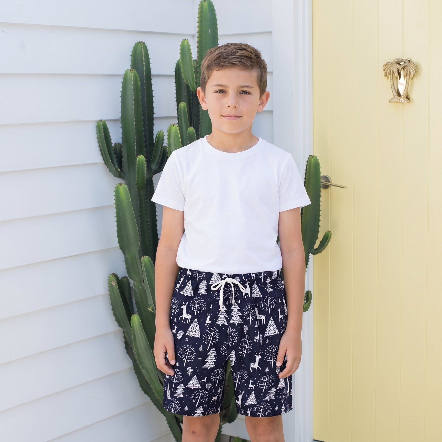 A young boy stands outdoors in front of a white house wall and a pale yellow door, wearing a plain white short-sleeve T-shirt and navy shorts with a white tree print and drawstring.
