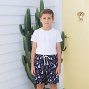 A young boy stands outdoors in front of a white house wall and a pale yellow door, wearing a plain white short-sleeve T-shirt and navy shorts with a white tree print and drawstring.