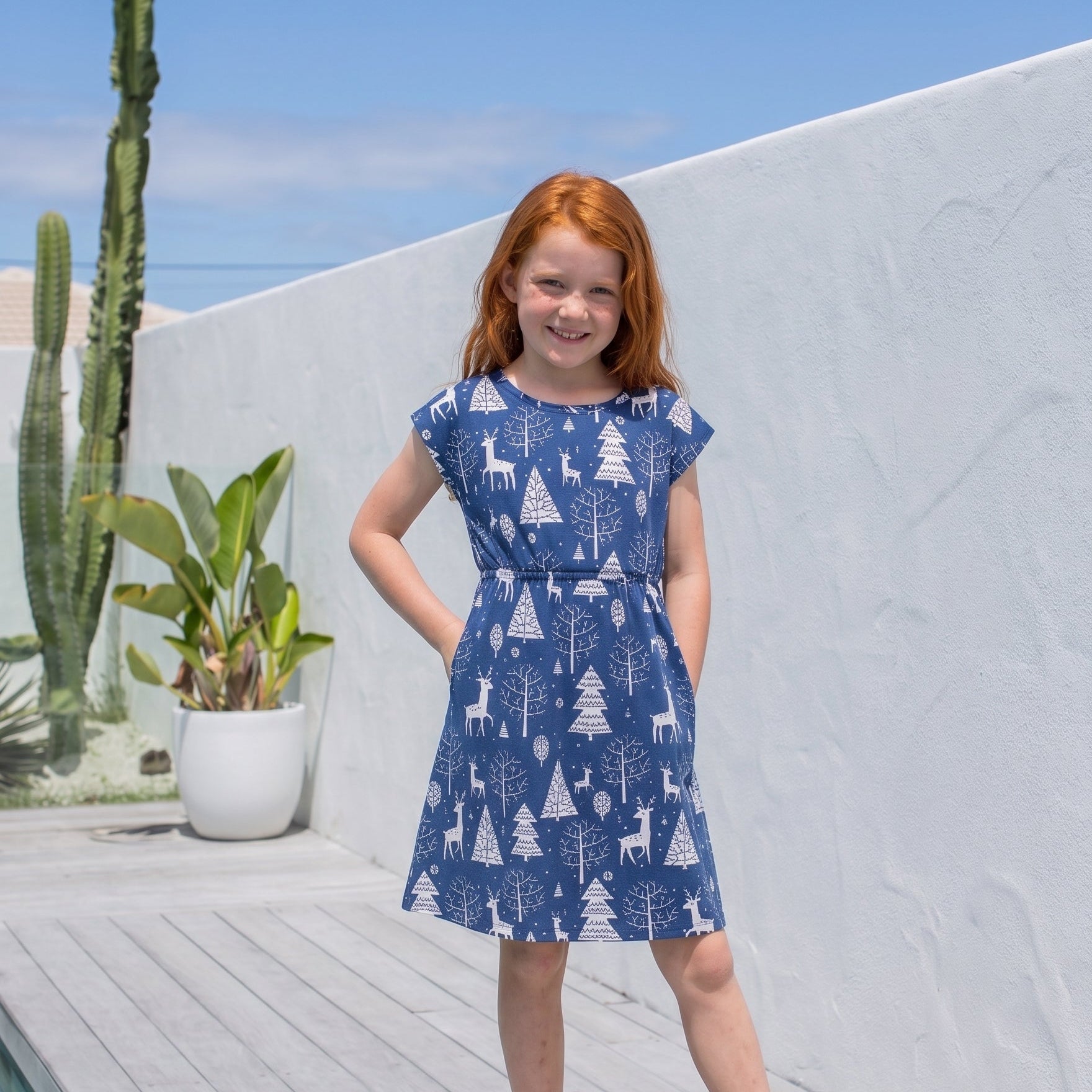 A young girl with red hair stands barefoot on a wooden deck wearing a blue sleeveless dress with a white tree and deer pattern, posing outdoors against a white wall and blue sky.