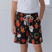 A young girl stands in a bright white room wearing black holiday-themed shorts with a gingerbread man, Santa Claus, and Christmas tree pattern, paired with a white t-shirt and beige shoes.