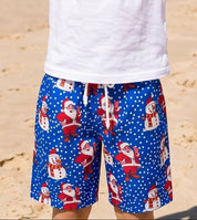 A young boy stands on a sandy beach with the ocean and blue sky behind him, balancing a pineapple on his head. He is wearing a plain white t-shirt and blue patterned shorts with Santa Claus designs.