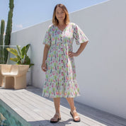 A woman wearing a short-sleeve tiered midi dress with a colorful floral print, standing on a wooden deck next to a pool with a white wall and cacti in the background.