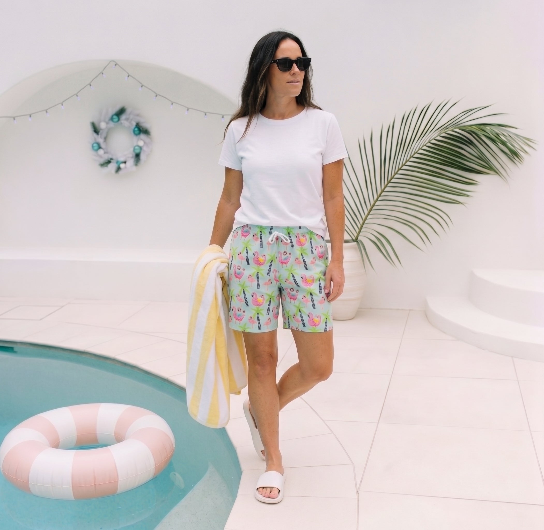 A woman wearing a white t-shirt and colorful tropical-print shorts walks beside a pool with a striped towel, in a bright, modern outdoor setting.