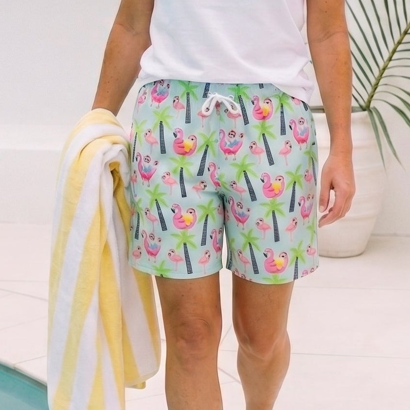 A woman walking beside a pool wearing a white t-shirt and colorful tropical print swim shorts, holding a yellow and white striped towel.