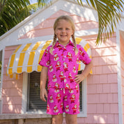 A young girl with blonde braids stands on a wooden deck in front of a pink house with a yellow and white striped awning, wearing a bright pink short-sleeve collared shirt and matching shorts set with a Santa Claus gift pattern.