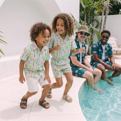 A family of four wearing matching tropical print swim shirts and shorts sets poses by a swimming pool. Two children are standing and smiling on the left, while two adults sit on the pool deck on the right.