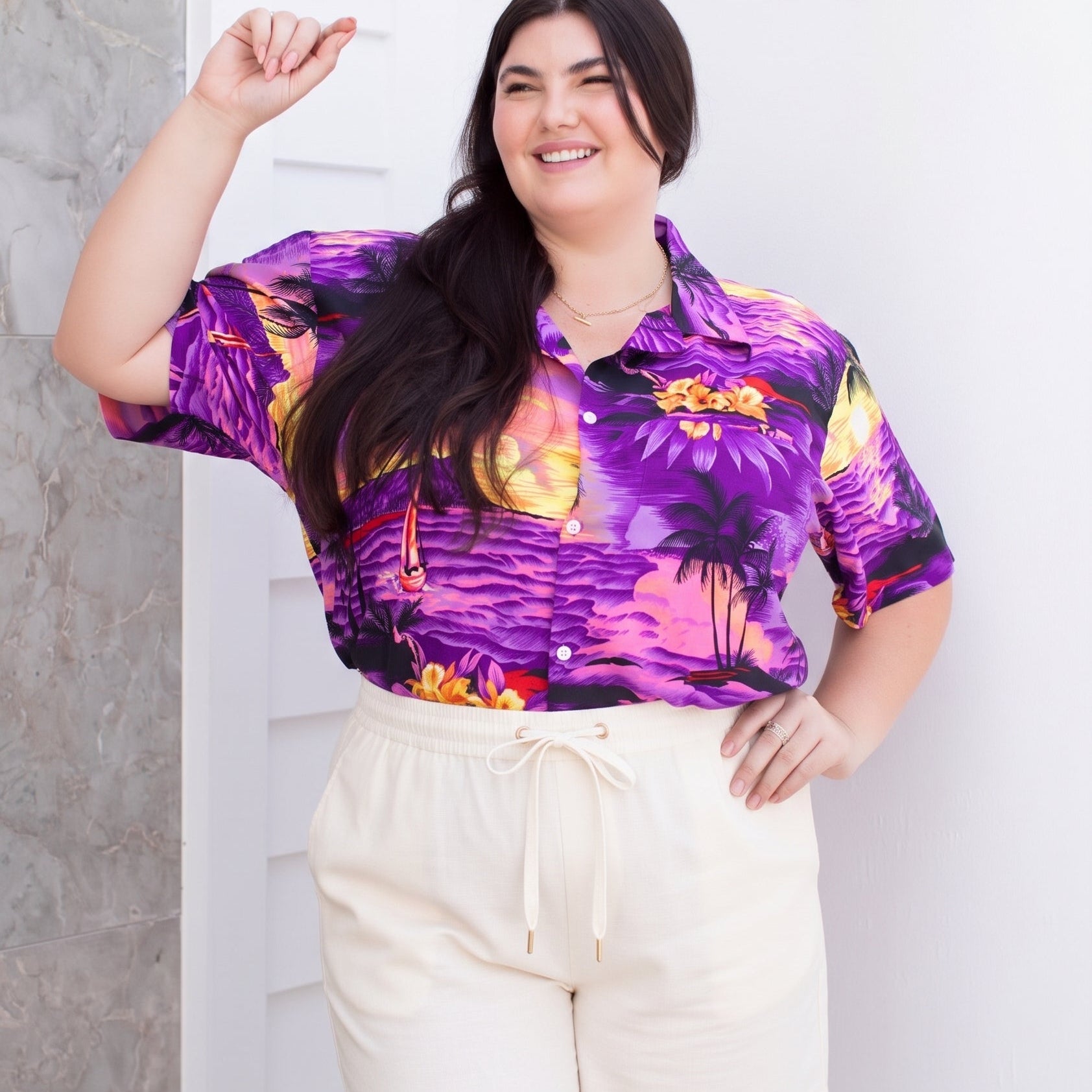 A woman modeling a short-sleeve button-up shirt with a vibrant purple, pink, and yellow tropical sunset print, paired with cream-colored drawstring shorts, standing against a white paneled wall.