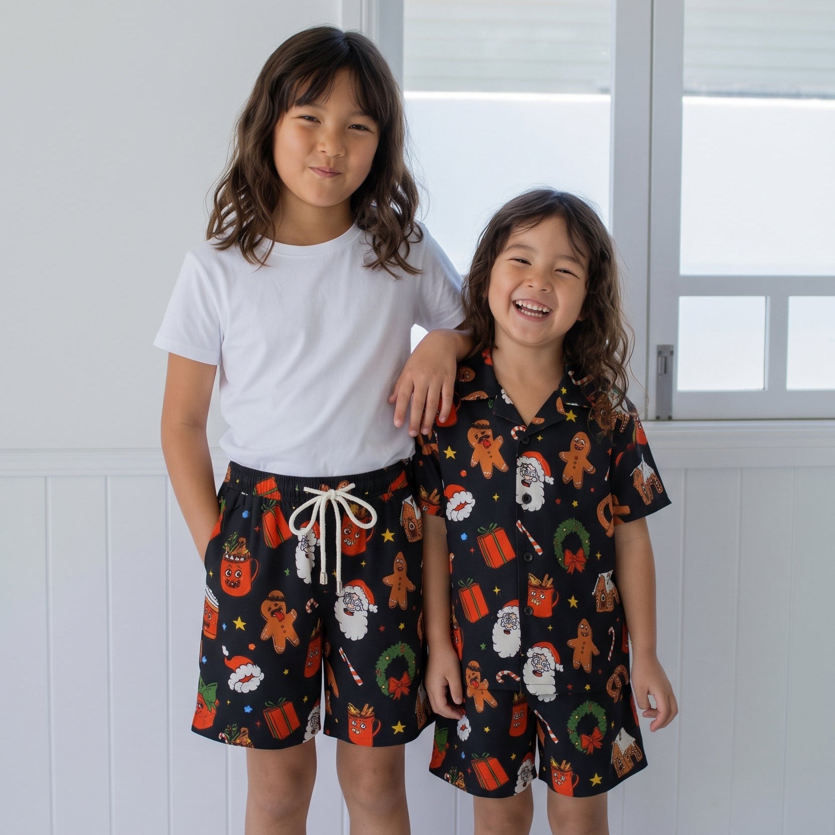 Two young girls standing indoors wearing matching dark holiday-themed shorts and a short-sleeve top set. The set features a black background with a colorful print of gingerbread men, Santa Claus, wreaths, and gifts.