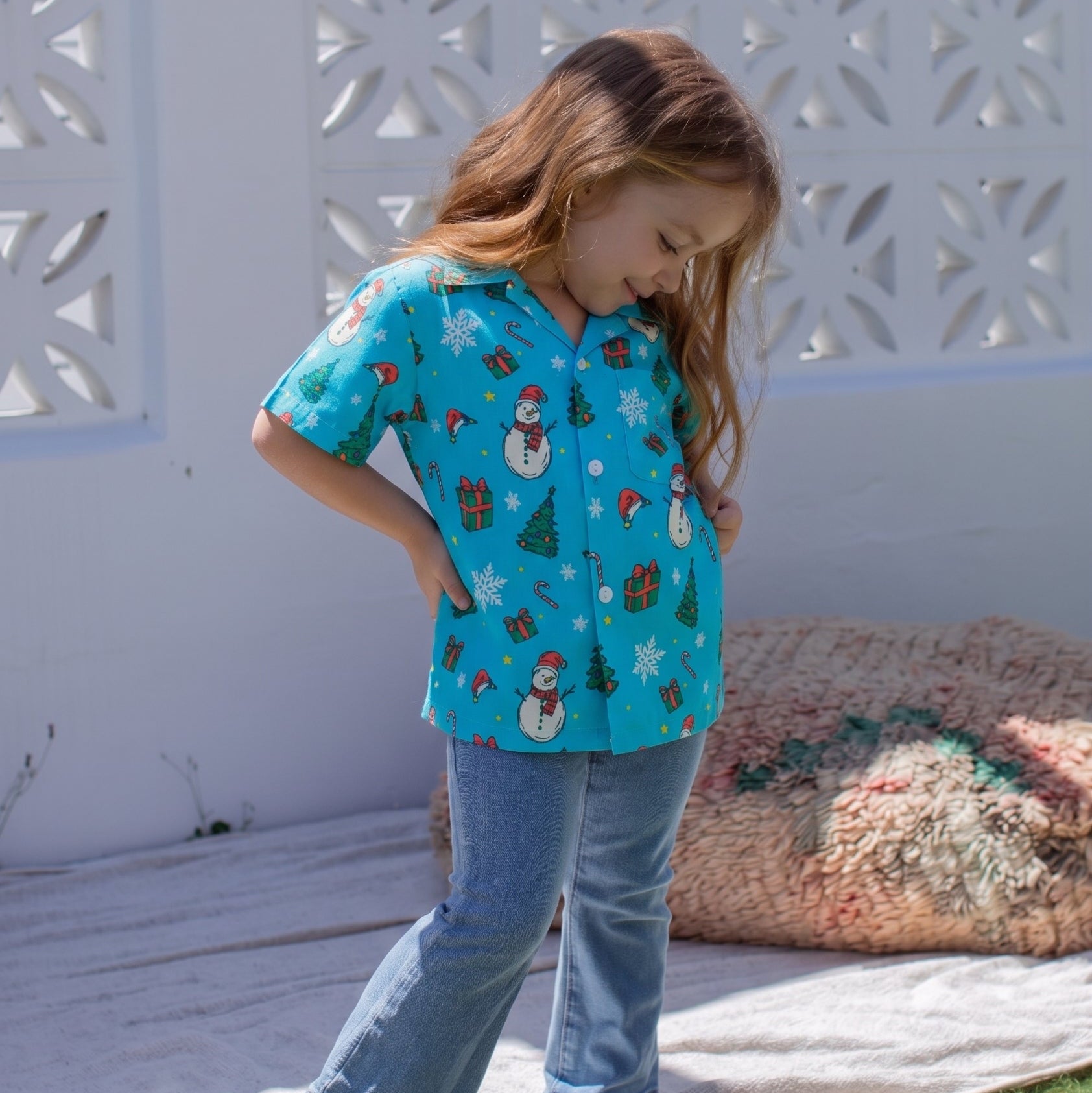 A young girl with long light brown hair stands barefoot outdoors on a white blanket, wearing a turquoise short-sleeve button-up shirt with a Christmas print and light blue jeans. The background features a white decorative lattice screen and a textured beige cushion.