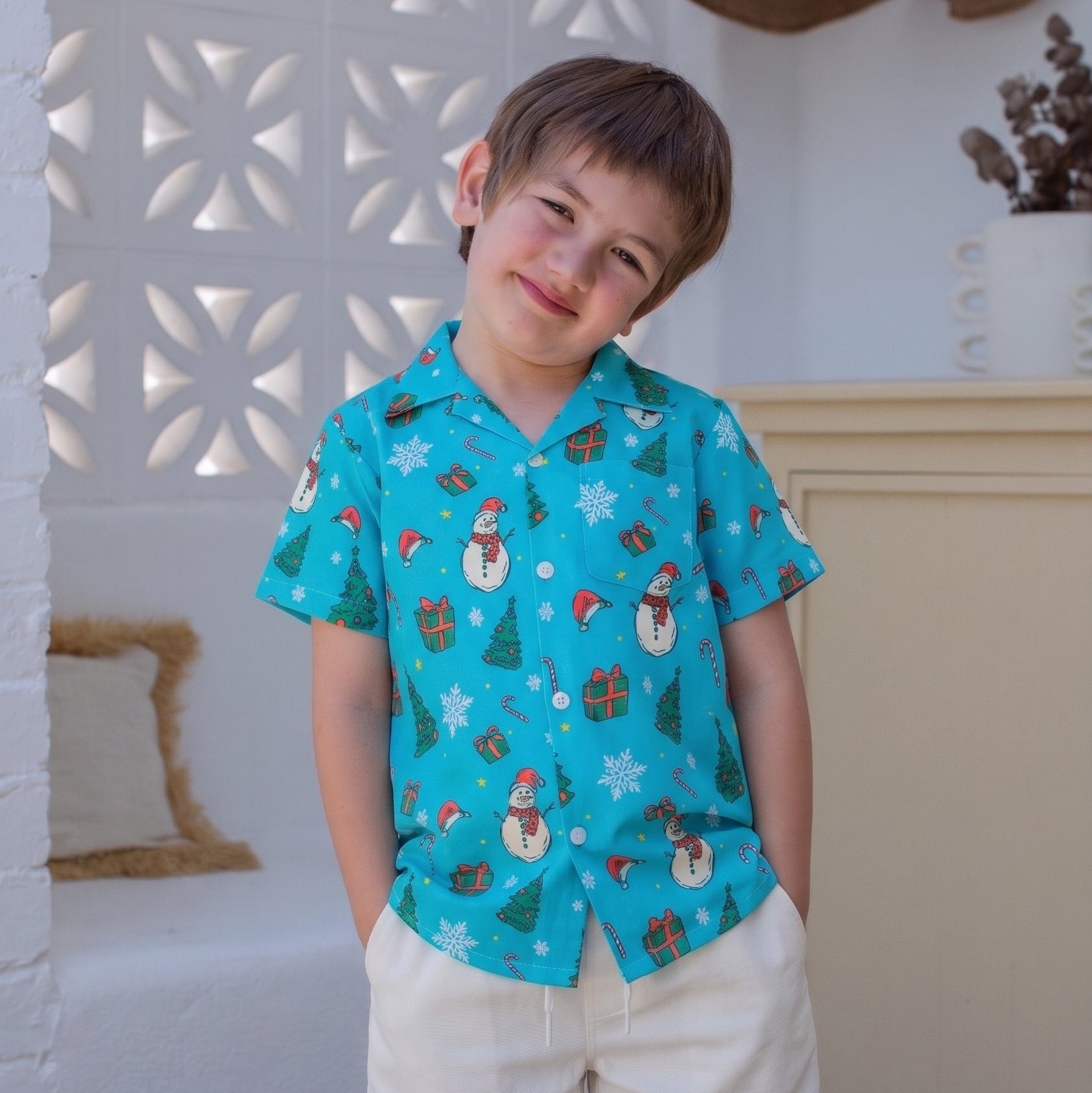 A young boy wearing a turquoise short-sleeve button-up shirt with a Christmas print of snowmen, trees, and gifts, paired with cream shorts, standing in a bright, modern room.