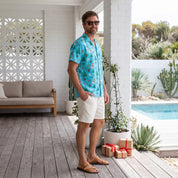 A man wearing a turquoise short-sleeve button-up shirt with a red and yellow tropical flamingo print, white shorts, and brown sandals stands on a wooden deck next to a swimming pool.