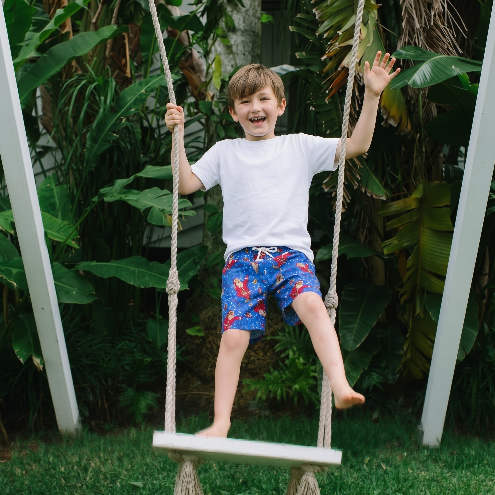 A young boy smiling and waving while sitting on a rope swing in a backyard with lush green tropical plants. He is wearing a plain white t-shirt and blue patterned swim trunks.