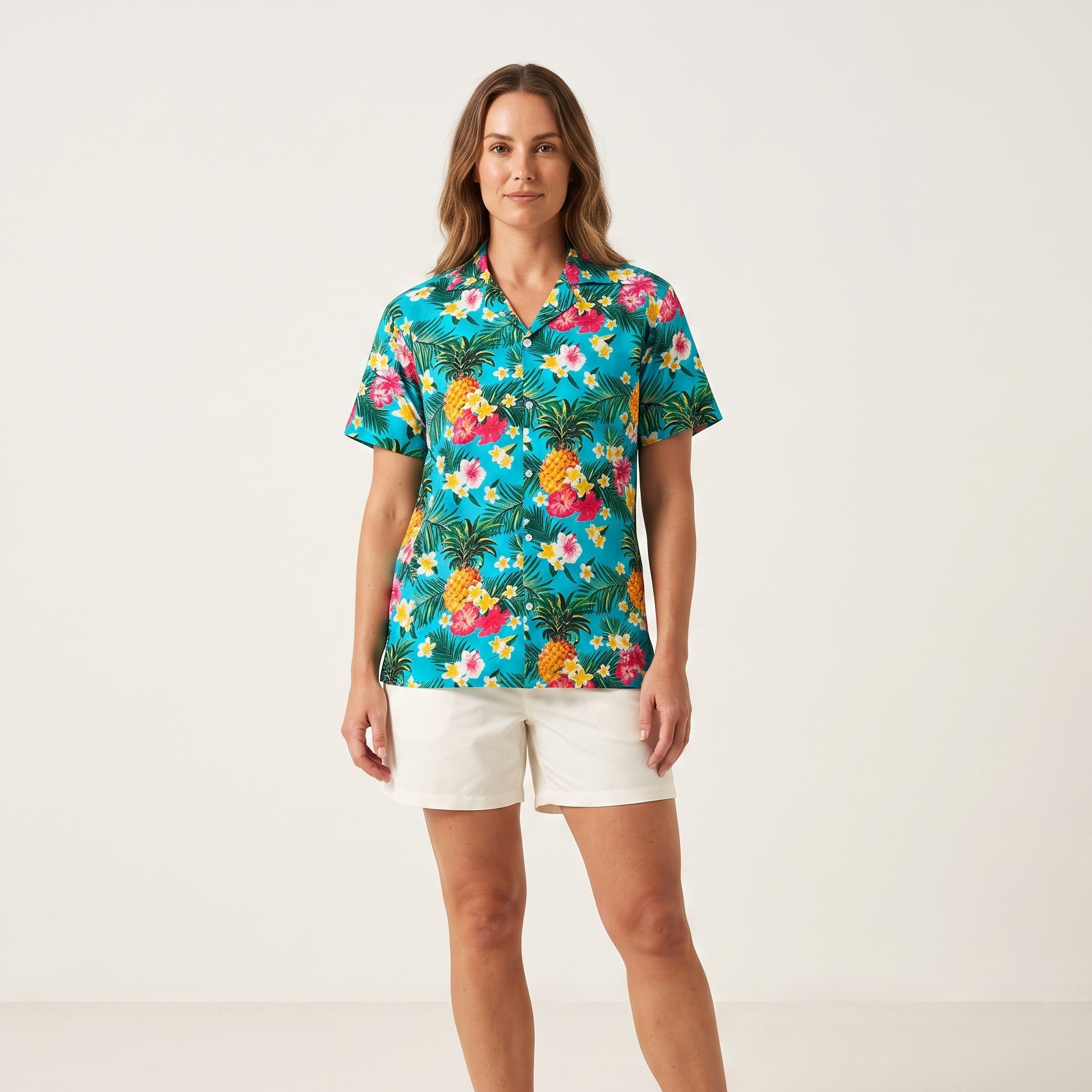 A woman stands barefoot against a clean off-white studio background, wearing a short-sleeve collared button-up shirt with a colorful tropical floral and pineapple print, paired with white shorts.