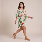 A woman modeling a tropical print kaftan dress with a tie waist, walking in a studio setting against a solid beige background.
