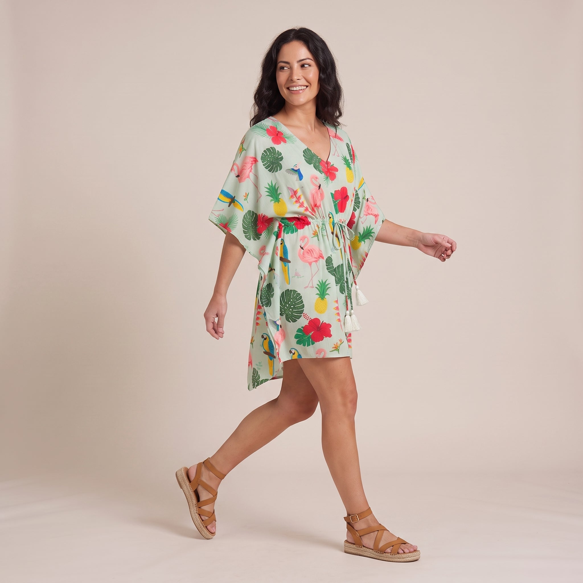 A woman modeling a tropical print kaftan dress with a tie waist, walking in a studio setting against a solid beige background.