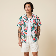 A man modeling a white short-sleeve button-up shirt with a vibrant tropical print of pink hibiscus flowers, green palm leaves, and orange koi fish, paired with off-white shorts against a solid beige background.