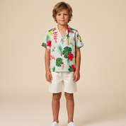 A young boy stands against a solid beige studio background wearing a short-sleeve tropical print button-up shirt, white shorts, and white sneakers.