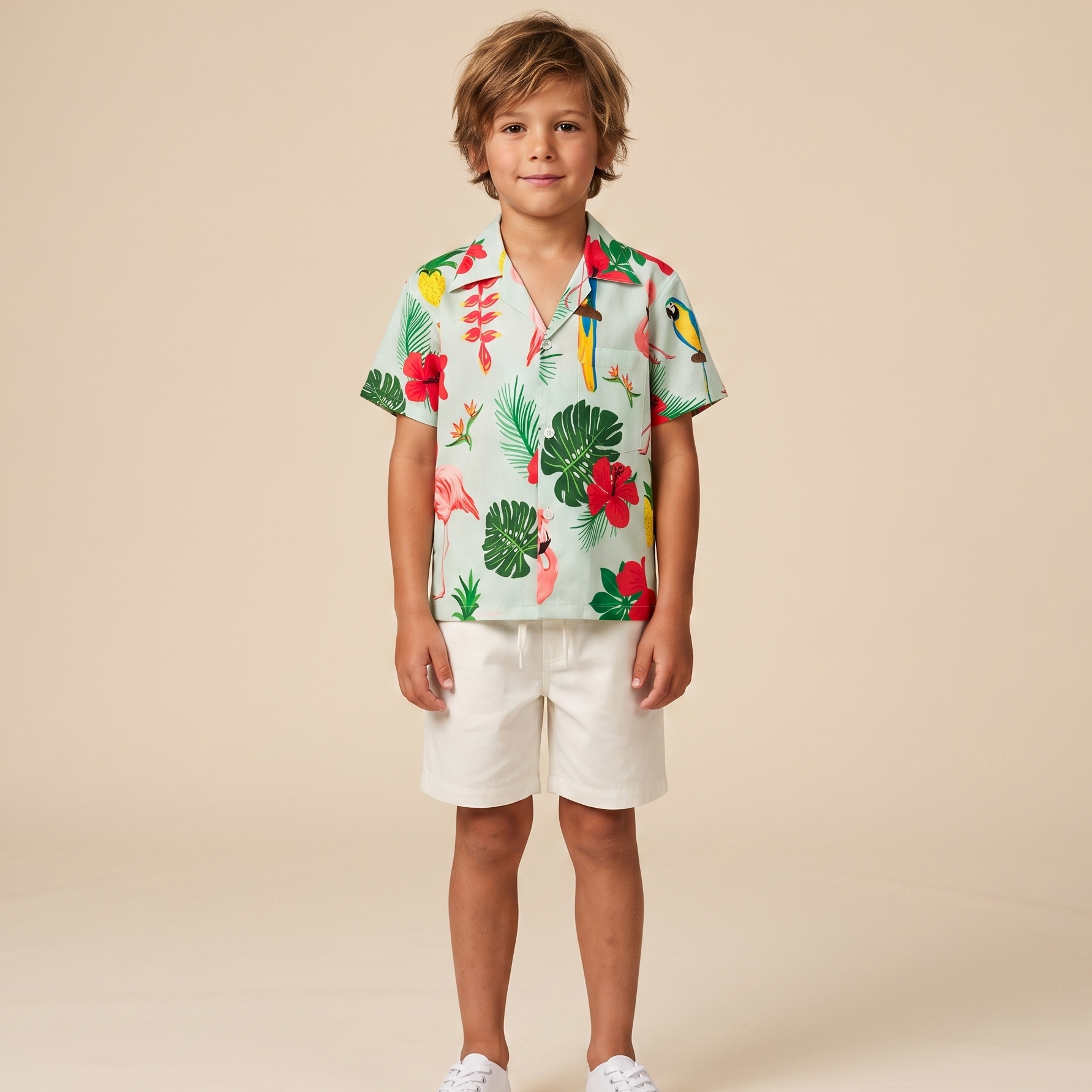 A young boy stands against a solid beige studio background wearing a short-sleeve tropical print button-up shirt, white shorts, and white sneakers.