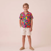 A young boy stands against a solid pale pink studio background wearing a short-sleeve button-up shirt with a vibrant multicolor abstract floral pattern, off-white shorts, and tan slip-on shoes.