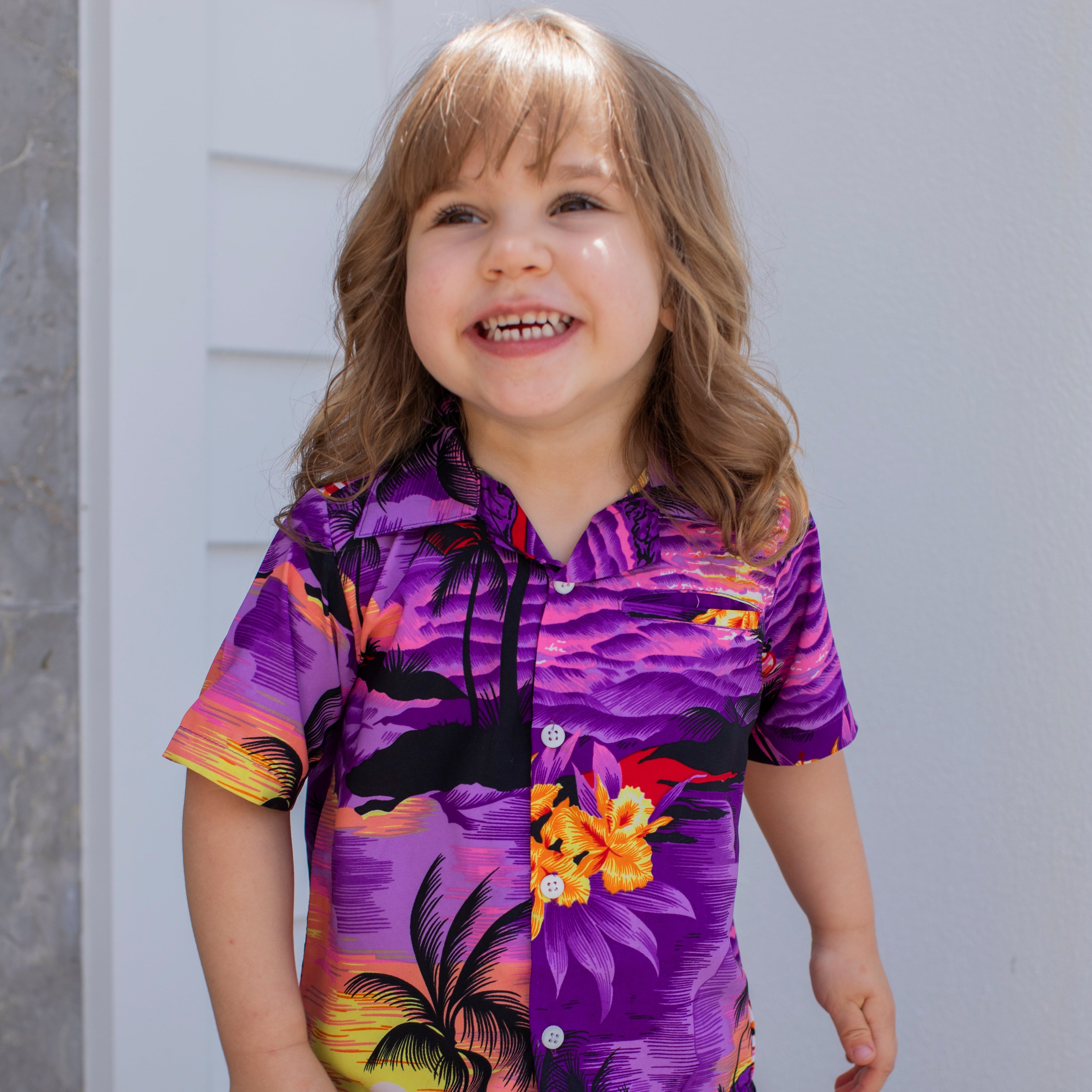 A smiling young child with long light brown hair showcases the Sunset Purple Kids Shirt, which features a vibrant design of palm trees and flowers, while standing outdoors in radiant sunlight.