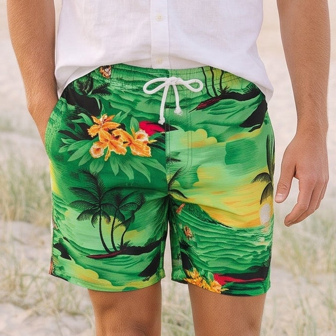 A man walking on a sandy beach wearing green tropical print swim trunks and a white short-sleeve button-down shirt.