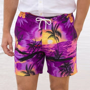 A man walking on a sandy beach wearing a white short-sleeve button-up shirt and purple tropical print swim trunks.