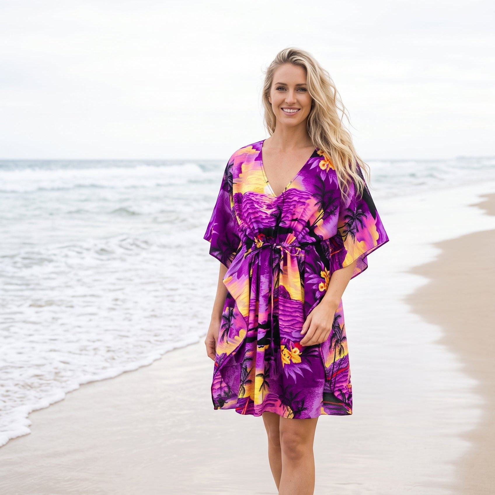 A woman stands barefoot on a sandy beach wearing a short, loose-fitting purple tropical print dress with yellow, orange, and black floral motifs.