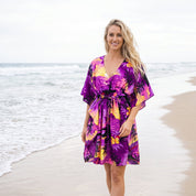 A woman stands barefoot on a sandy beach wearing a short, loose-fitting purple tropical print dress with yellow, orange, and black floral motifs.